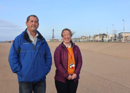 Image of a man standing next to a woman. He is wearing a blue coat and the woman is wearing an orange jumper with a purple coat. In the background you can see the pier and the sky is clear and blue.