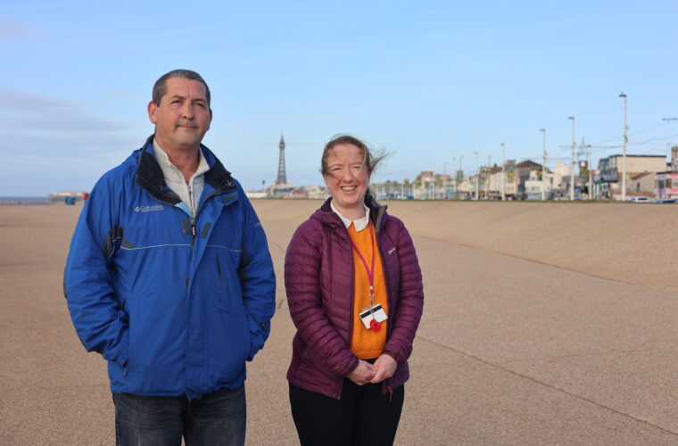Image of a man standing next to a woman. He is wearing a blue coat and the woman is wearing an orange jumper with a purple coat. In the background you can see the pier and the sky is clear and blue.